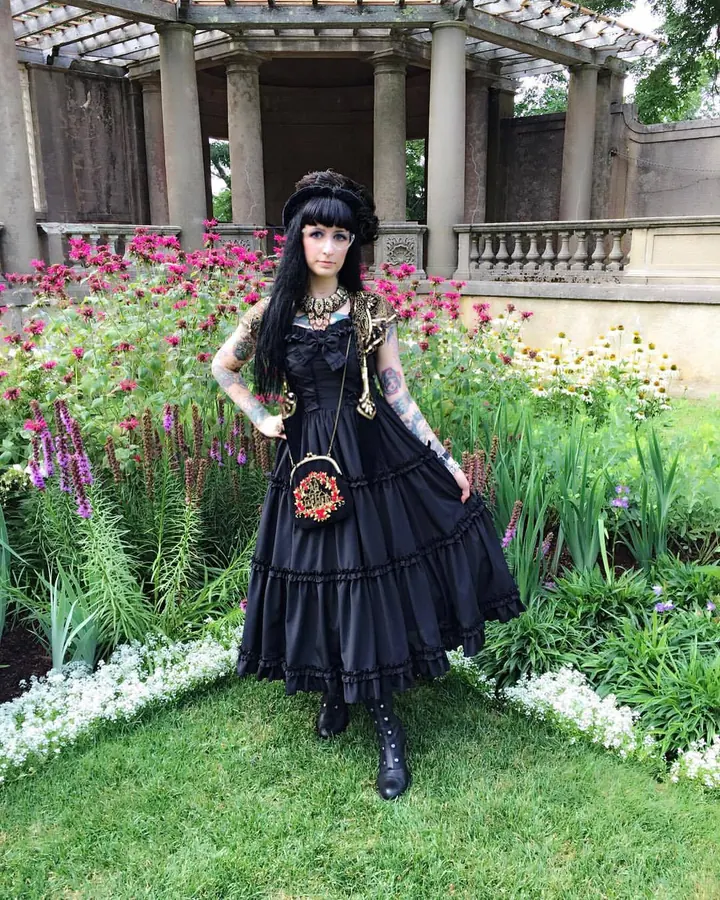 Femme aux cheveux noirs longs, vêtue d'une robe noire gothique avec des manches transparentes, debout dans un jardin fleuri, mains sur les hanches.
