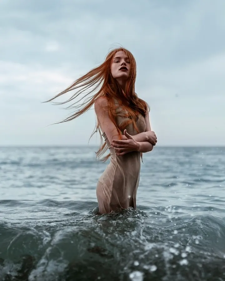 Femme rousse aux yeux marrons, portant un haut noir, regardant directement l'objectif avec une expression posée, sur fond sombre.