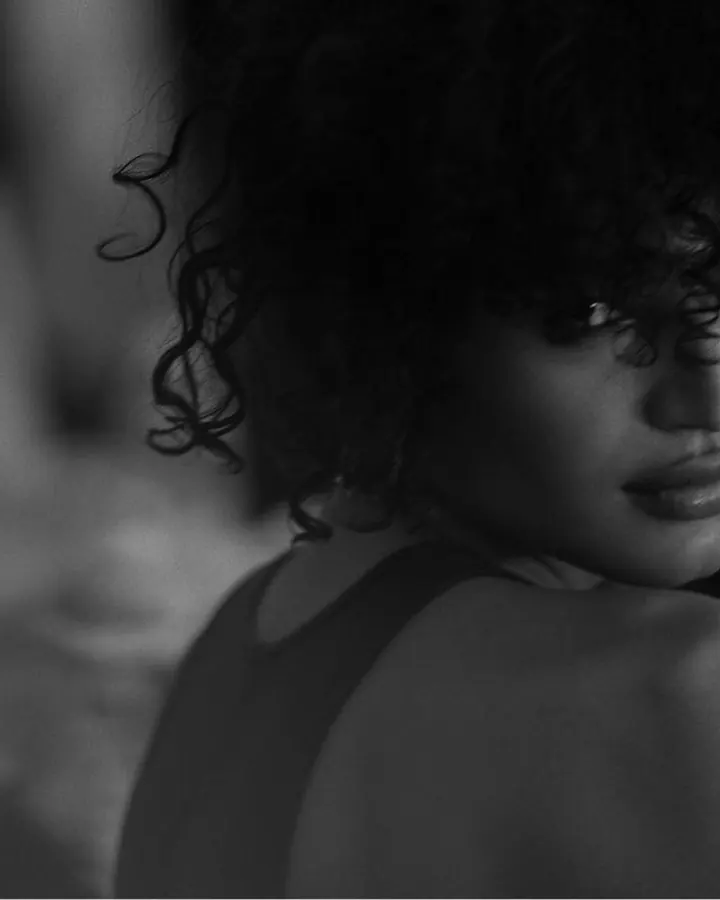 Photo en noir et blanc d’une femme aux cheveux bouclés, regardant par-dessus son épaule, vêtue d’un haut sans manches
