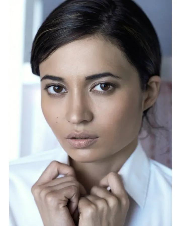 Femme brune en chemise blanche, regard intense, mains serrées autour du col, ambiance sobre et professionnelle