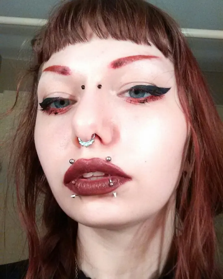 Visage de femme aux cheveux roux, yeux bleus, maquillage sombre et piercings multiples, regard intense