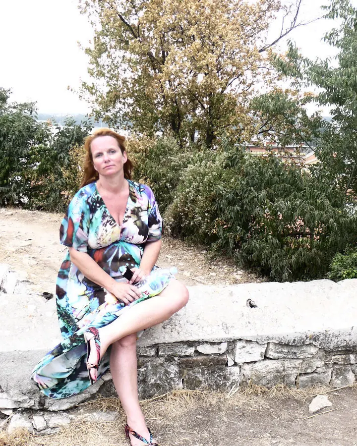 Femme aux cheveux blonds, portant un haut jaune doré et une jupe courte, debout devant un buisson fleuri.
