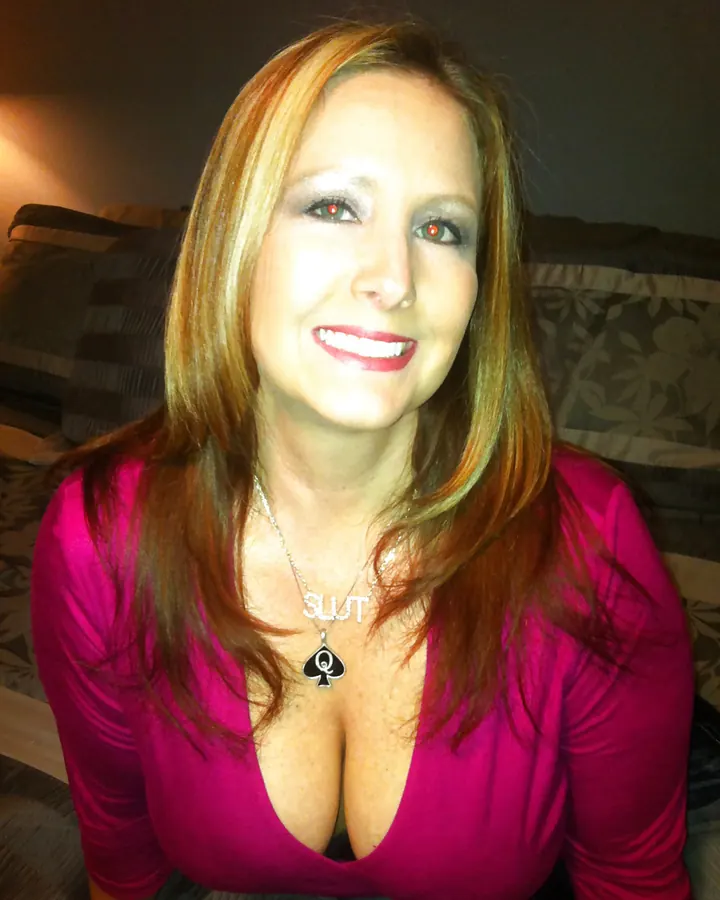 Femme aux cheveux roux, souriante, portant un haut rose foncé, collier avec pendentif "SLUT", dans un salon.