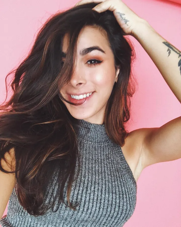 Femme aux cheveux châtains souriante devant un fond rose, portant un haut gris sans manches, main dans les cheveux.