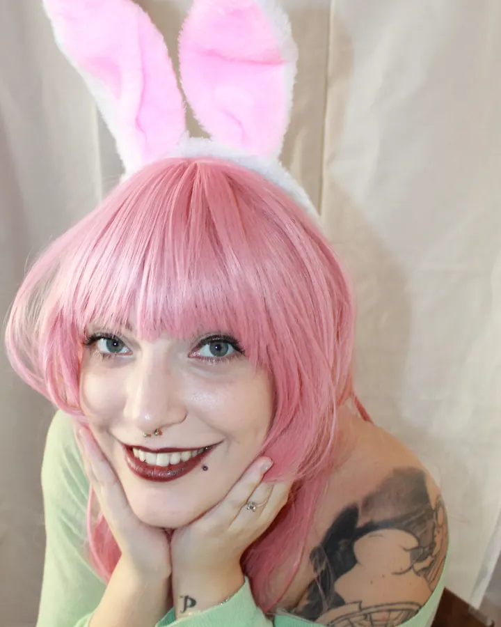 Femme aux cheveux roses, portant des oreilles de lapin, un sourire large, les mains sur les joues, avec un tatouage visible sur l'épaule.