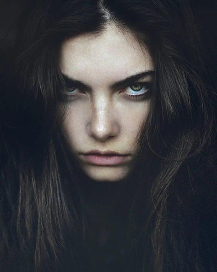 Femme aux cheveux roux, vêtue d'une robe noire, debout sur un trottoir enneigé, portant un chapeau noir et une veste noire avec col en fourrure.