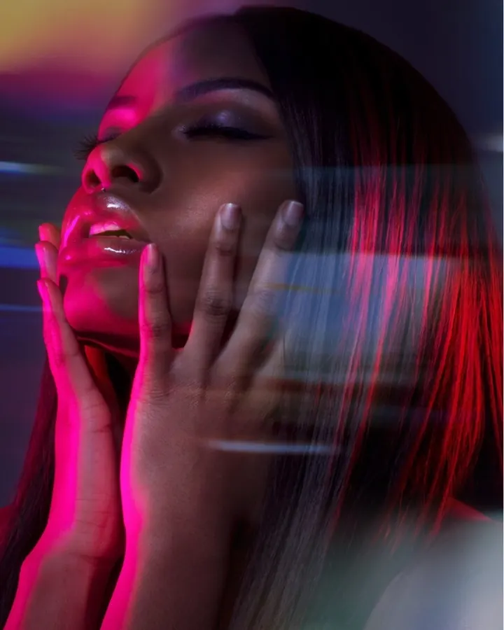 Femme européenne aux cheveux roux portant un haut noir et un jean, posant près d'un lit avec un tête de lit en velours noir.