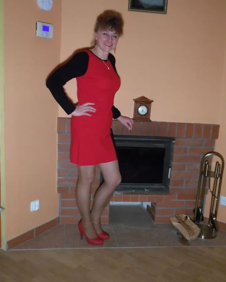 Femme souriante en robe rouge courte, mains sur les hanches, devant une cheminée en briques