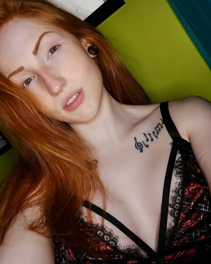 Femme aux cheveux roux, vêtue d'un haut noir, tenant sa main près de son visage, tatouage visible sur le bras.