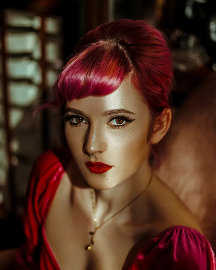 Femme aux cheveux roses vifs, maquillage soigné, robe rouge satinée, assise dans un fauteuil sombre, regard fixe et intense.